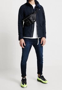 Marinjacka med hög krage, svart midjeväska, vit t-shirt, mörka jeans och svarta sneakers med neongröna accenter.