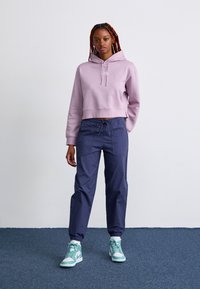 Haut à capuche cropped lilas avec détails de cordon, associé à un pantalon à taille élastique bleu marine et des baskets montantes teal. Textures lisses tout au long.