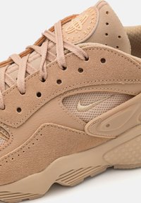 Beige Nike-sneaker i närbild som visar läder- och meshtexturer, perforeringar, snören, Nike swoosh-logotyp och detaljerad sula.