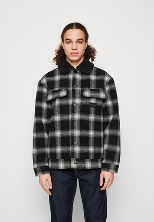 Chaqueta de cuadros en blanco y negro con cierre de botones, dos bolsillos en el pecho y un cuello texturizado en negro. La tela tiene una apariencia suave y similar a la lana.