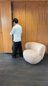 Görbült, bézs színű kárpitozott szék puha textúrával, sötét fa padlón áll. A háttérben fából készült szekrényajtók félig nyitva vannak.