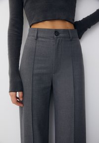 Vrouw in hoge, brede grijze broek met een cropped, donkergrijze, lange mouwen geribbelde trui met duimgaten.