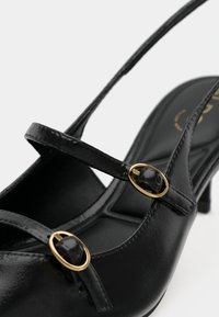 Chaussure à talon haut en cuir noir avec deux fines brides ornées de boucles ovales dorées, présentée sur un fond blanc uni.