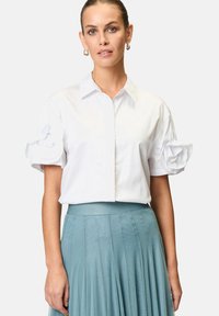 Camicia bianca a maniche corte con bottoni sul davanti e dettagli in rosa di tessuto oversize sulle maniche. Abbinata a una gonna plissettata color teal per un contrasto.