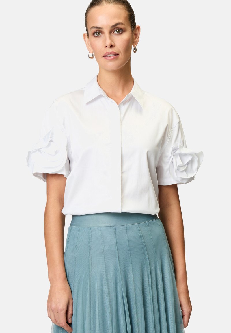 Camicia bianca a maniche corte con bottoni sul davanti e dettagli in rosa di tessuto oversize sulle maniche. Abbinata a una gonna plissettata color teal per un contrasto.