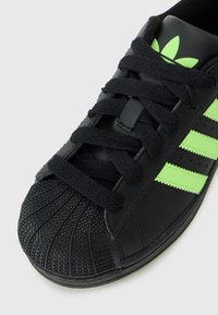Baskets noires avec une coque texturée, lacets noirs et rayures et logo Adidas vert fluo sur la languette, sur fond blanc.