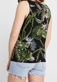 Top senza maniche nero con vivaci stampe botaniche verdi e rosa, caratterizzato da un piccolo taglio a forma di chiave nella parte posteriore, abbinato a pantaloncini di denim.