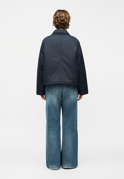 Άτομο με κοντά σγουρά μαλλιά φοράει σκούρο μπουφάν με επένδυση και φαρδιά μπλε τζιν παντελόνια, στέκεται με την πλάτη προς το φωτεινό απλό φόντο.