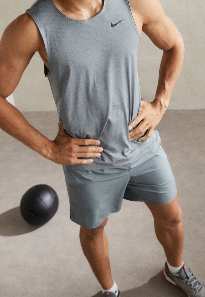 Graues athletisches Tanktop mit schwarzem Logo, kombiniert mit passenden grauen Shorts. Glatte Textur und taillierte Passform, mit Händen auf den Hüften. Schwarzer Trainingsball in der Nähe.