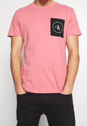 Mann trägt ein rosa T-Shirt mit kurzen Ärmeln und einer schwarzen Brusttasche, die ein weißes, rundes "Calvin Klein Jeans"-Logo zeigt, sowie schwarze Hosen.