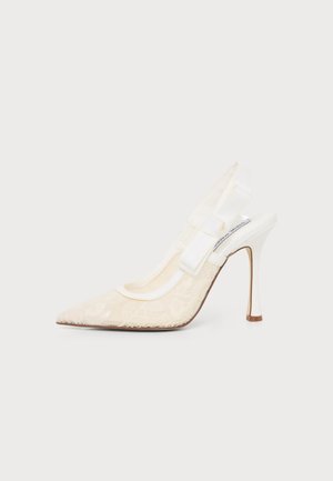 Chaussure à talon haut blanche en dentelle avec bout pointu et nœud transparent sur la bride, sur fond uni.