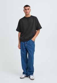 Sweatshirt nero oversize con maniche corte, abbinato a jeans in denim blu larghi e sneakers bianche con strisce nere.