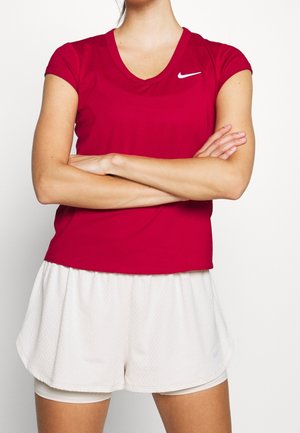 Personne les bras croisés portant un t-shirt rouge Nike à manches courtes et un short de sport blanc sur un fond blanc uni.