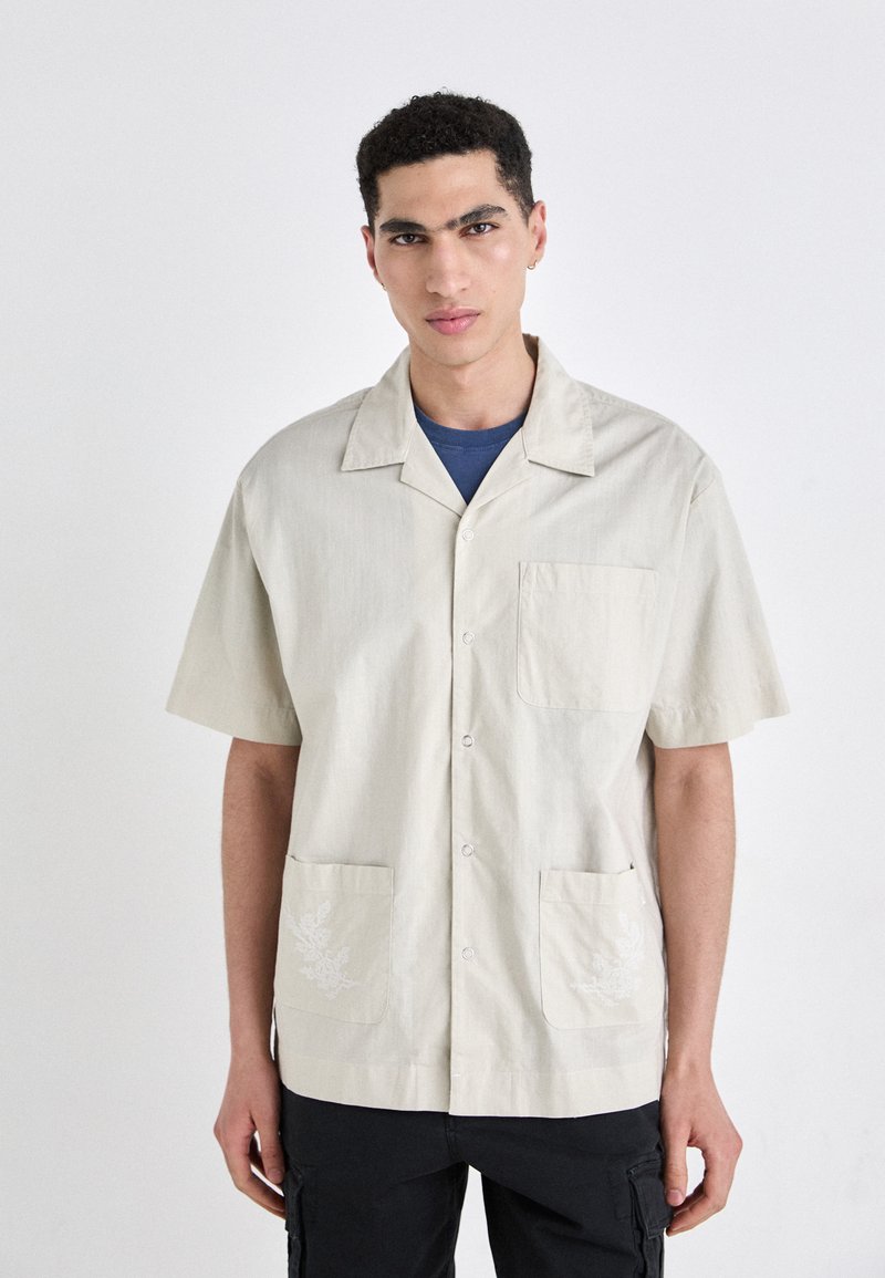 Camisa de manga corta en beige claro con cuello, bolsillo en el pecho y dos bolsillos frontales que presentan un sutil bordado floral.