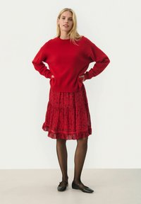 Rode gebreide trui met lange mouwen, gecombineerd met een gerimpelde rode bloemenrok. Zwarte platte schoenen en doorschijnende zwarte panty’s maken de outfit compleet.