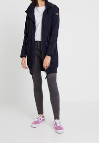 Granatowa długa kurtka z kapturem, zamek błyskawiczny i elastyczne detale. Połączona z szarymi teksturowanymi legginsami i różowymi/białymi trampkami.