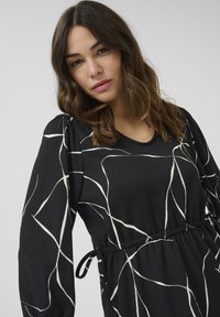 Robe noire avec un motif abstrait blanc, manches longues, épaules froncées et taille élastique avec un lien décoratif. Texture lisse.