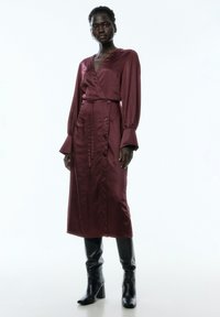 Satinburgunder-Wickelkleid mit langen Ärmeln, tailliertem Schnitt, knöpfbarem Detail vorne und fließendem Design, das bis zur Mitte der Wade reicht. Kombiniert mit schwarzen Stiefeletten.