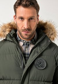 Homme portant une veste d'hiver matelassée vert foncé avec une capuche en fourrure, et une chemise à carreaux boutonnée en dessous, regardant droit devant lui.