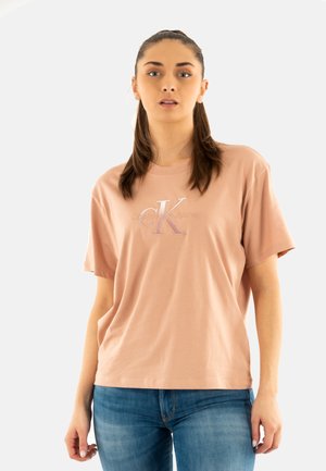 Camiseta de algodón rosa con mangas cortas, que presenta un logo "CK" texturizado en púrpura claro, corte holgado y cuello redondo. Combinada con jeans azules.