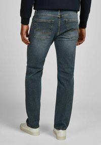 Homme portant un jean bleu délavé et des baskets blanches, se tenant avec les mains détendues le long du corps, devant un fond clair et uni.