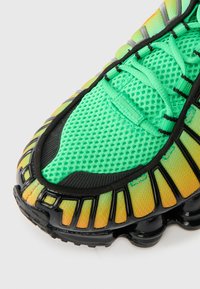 Bota atlética com biqueira, parte superior em malha verde, lados em degradê amarelo-laranja, sola preta com design segmentado flexível e atacadores verdes.