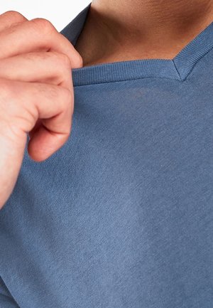 Ręka ciągnąca kołnierz niebieskiej koszulki z dekoltem w serek, ukazująca linię szyi i górną część klatki piersiowej.