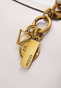 Brelok z mosiądzu z geometrycznymi i okrągłymi zawieszkami, w tym jedna prostokątna zawieszka z wygrawerowanym napisem "GUESS" na białej fakturowanej powierzchni.