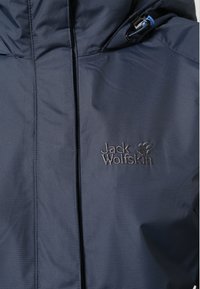 Granatowa wodoodporna kurtka z wysokim kołnierzem, przednim zamkiem błyskawicznym oraz haftowanym logo. Tkanina ma subtelną fakturę i jest wytrzymała.