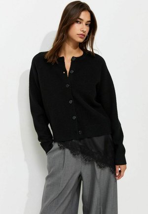Vrouw met lang bruin haar die een zwart vest met knopen draagt over een zwarte kanten top en losse grijze broek, staand tegen een witte achtergrond.