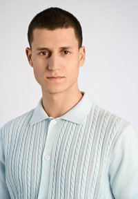 Vaaleansininen neulepolo, jossa on kaapelikuvioita, lyhyet hihat ja kaksi nappia kauluksessa. Silkin pehmeä pinta ja miellyttävä kangas.