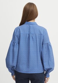 Blusa blu con un motivo geometrico ripetuto, maniche lunghe a sbuffo e colletto. Tessuto liscio con una finitura leggermente lucida.