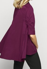 Blusa color prugna con orlo asimmetrico, maniche arrotolate e dettagli arricciati ai fianchi. Realizzata in un tessuto morbido, abbinata a pantaloni neri.
