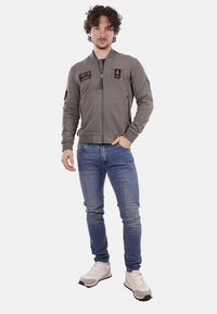 Giacca grigia con zip, dotata di due tasche frontali e toppe ricamate; indossata sopra jeans blu e sneakers beige. Tessuto morbido con polsini a costine.