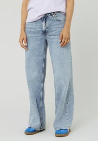 Jeans en denim bleu clair à larges jambes avec un look décoloré, cinq poches et des détails de couture visibles. Portés avec des chaussures bleues et une coupe décontractée.