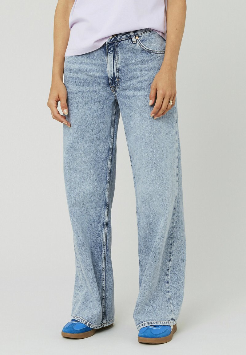 Jeans en denim bleu clair à larges jambes avec un look décoloré, cinq poches et des détails de couture visibles. Portés avec des chaussures bleues et une coupe décontractée.