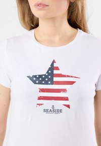 Witte t-shirt met een grote sterafbeelding met een verouderd ontwerp van de Amerikaanse vlag. Tekst luidt "SEASIDE NAUTICAL LEGACY" in marineblauw.