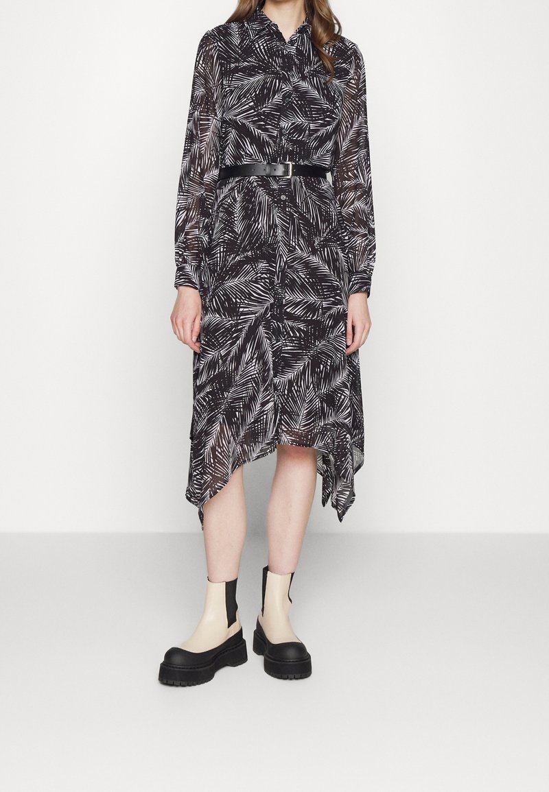 Femme portant une robe à manches longues à motif feuilles noir et blanc avec un ourlet asymétrique et une ceinture noire, associée à des bottes à plateforme beige et noire.