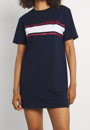 Kobieta w granatowej sukience w stylu T-shirt z krótkim rękawem, białymi i czerwonymi poziomymi paskami oraz logo „Hollister” na piersi.