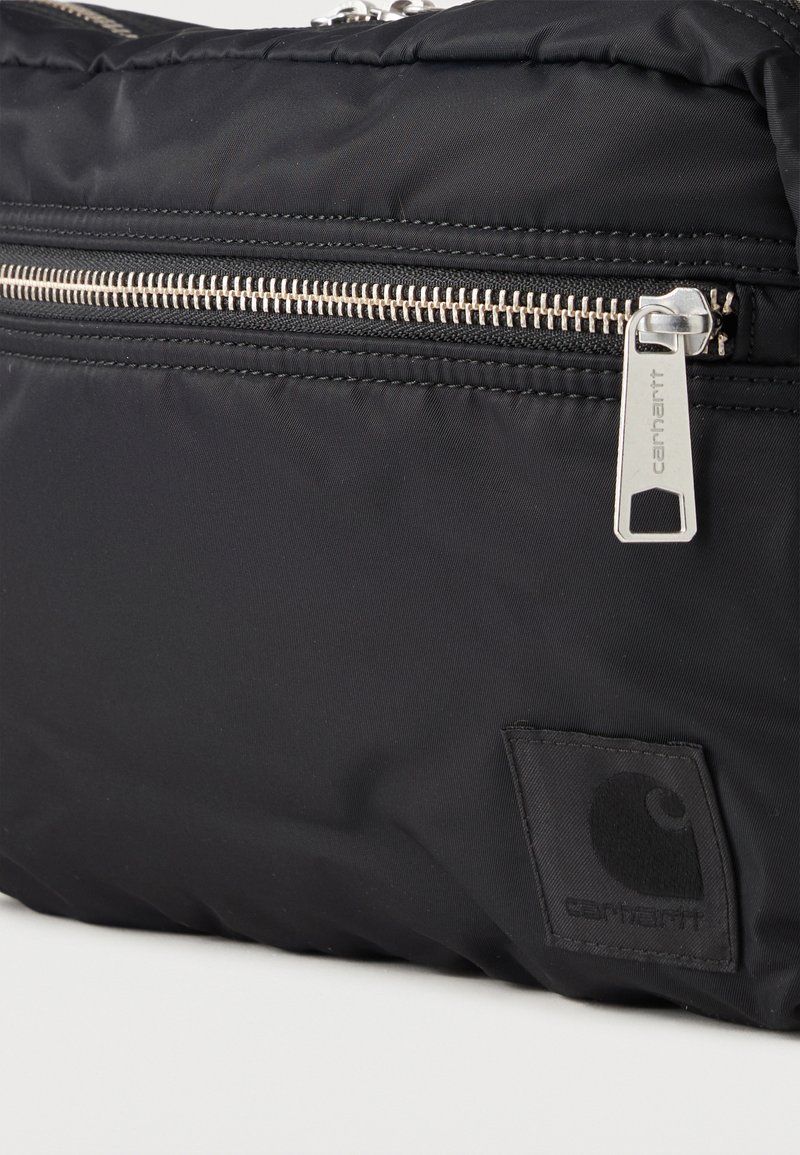 Sac en tissu noir avec fermeture éclair argentée horizontale visible et tirette en métal gravée «Carhartt» ainsi qu’un écusson logo cousu.