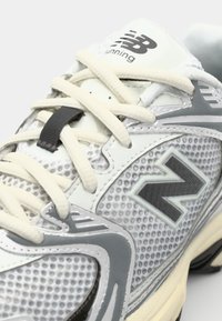 Scarpa da corsa New Balance grigia con tomaia in rete, lacci bianchi e dettagli neri. Presenta un prominente logo 'N' e una suola texturizzata.