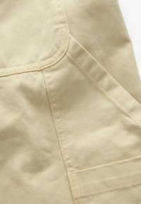 Tissu en coton beige avec une texture lisse, comportant une couture et une poche plaquée. Des accents de couture le long des bords sont visibles.