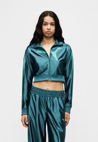 Γυναίκα που φοράει μπλε-πράσινο γυαλιστερό κοντό μπουφάν με φερμουάρ και ταιριαστό παντελόνι με το λογότυπο της Puma, στέκεται μπροστά σε ένα απλό φωτεινό φόντο.