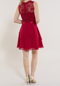 Robe rouge avec haut en dentelle et jupe en mousseline superposée ; taille cintrée avec un ruban en satin. Portée avec des chaussures à talons métalliques.