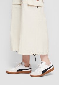 Baskets blanches avec des accents noirs, semelle en caoutchouc et dessus texturé. Lacets et branding subtils visibles ; associées à une jupe crème.