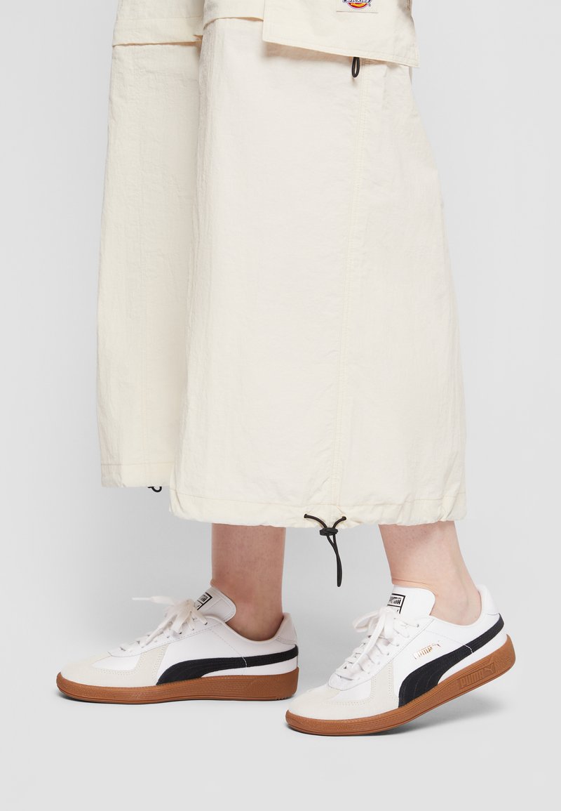 Baskets blanches avec des accents noirs, semelle en caoutchouc et dessus texturé. Lacets et branding subtils visibles ; associées à une jupe crème.
