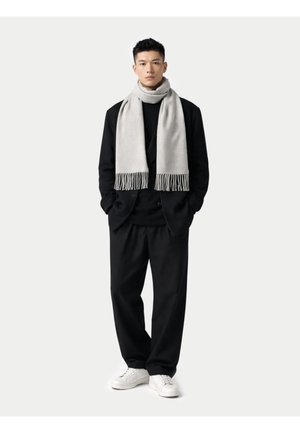 Mann steht mit den Händen in den Taschen, trägt einen schwarzen Anzug, ein schwarzes Hemd, einen hellgrauen Schal und weiße Turnschuhe vor einfarbigem Hintergrund.