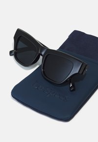 Mustat suorakulmaiset aurinkolasit tummilla linsseillä, jotka ovat tummansinisessä kankaisessa kotelossa, jossa on kohotettu teksti "Le Specs". Sileä muovipinnoite.