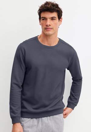 Junger Mann trägt einen dunkelgrauen Sweatshirt mit Rundhalsausschnitt und hellgraue Hose vor weißem Hintergrund.