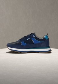 Αθλητικό παπούτσι σε ναυτικό μπλε και μαύρο με λευκή και πράσινη σόλα, μπλε λεπτομέρειες και σχέδιο με κορδόνια, πάνω σε φωτεινή επιφάνεια με γκρι φόντο.